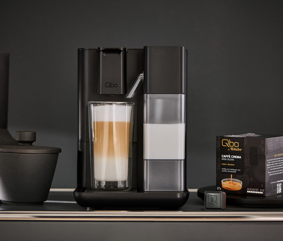 Ein Qbo MILK MASTER Velvet Black bereitet einen Latte Macchiato zu. Daneben stehen Kaffeekapseln und eine Tasse.