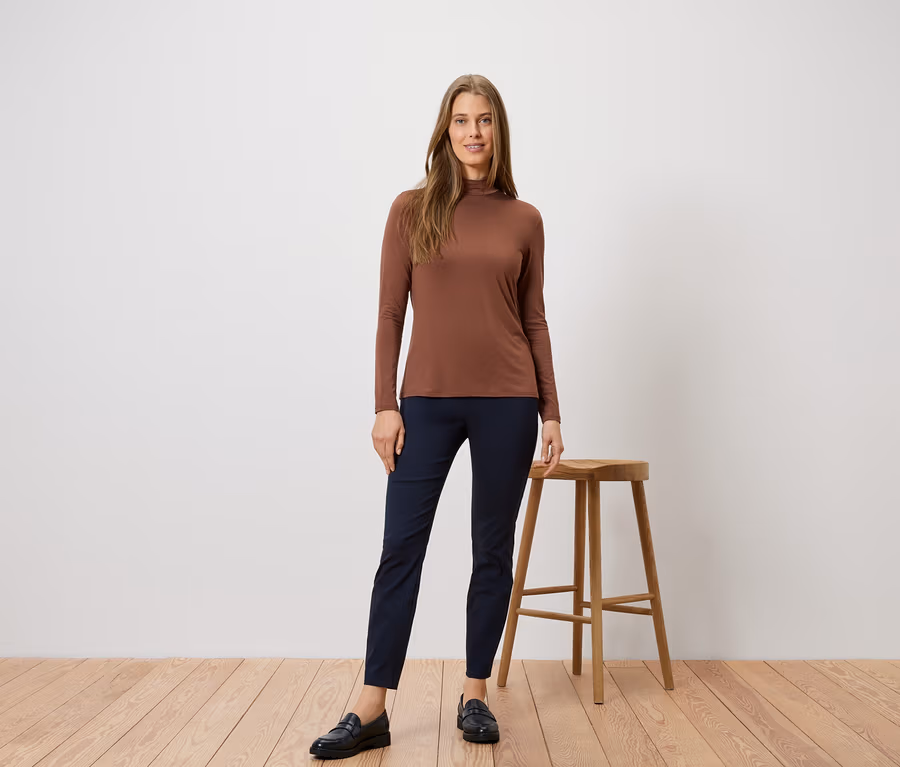Frau in braunem Rollkragenshirt und blauen Hosen posiert neben einem Hocker auf einem Holzboden.