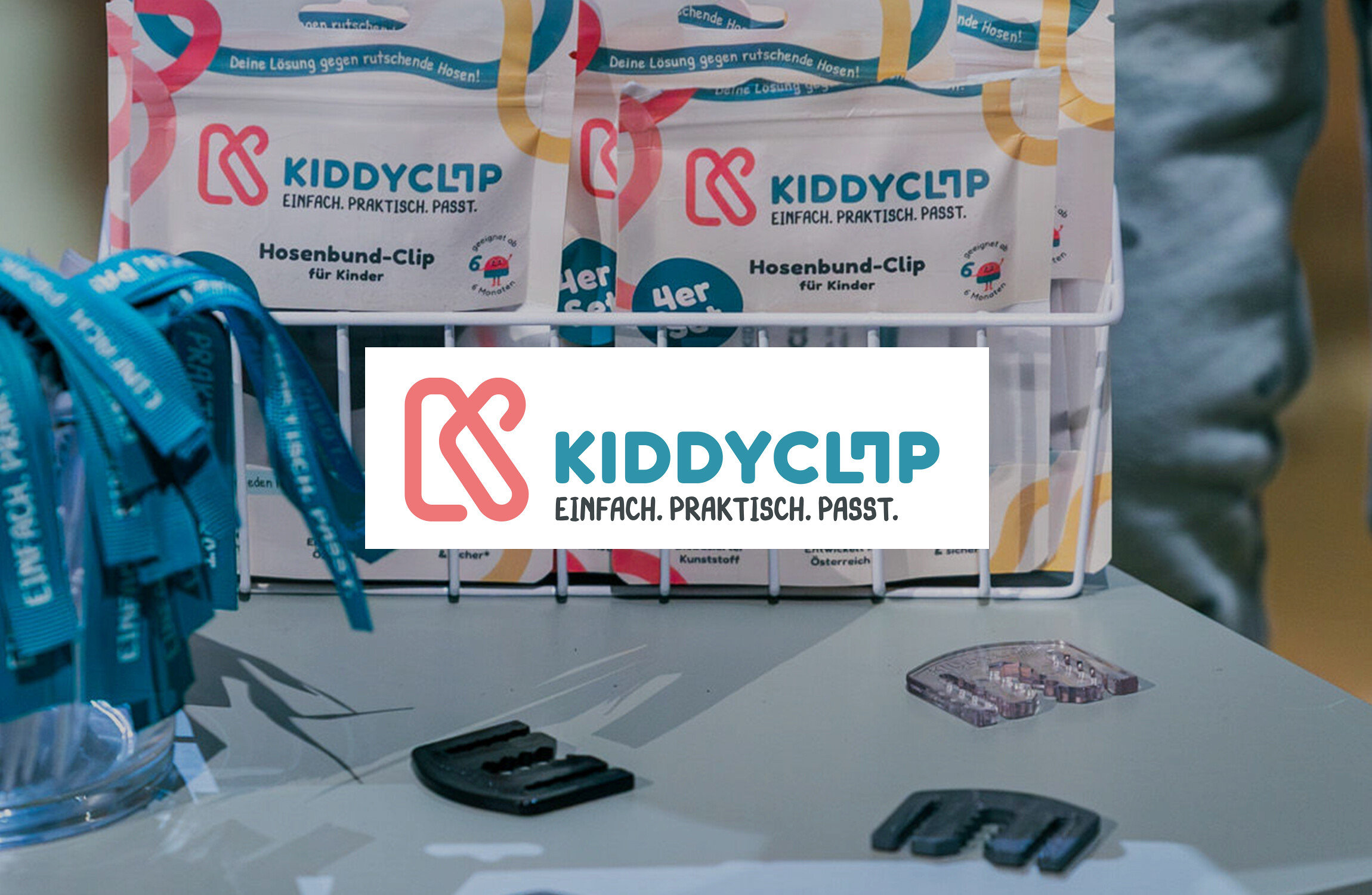 Auf einem Tisch stehen KiddyClips Hosenbund-Clips in Verpackungen und lose, daneben blaue Bänder in einem Glas.