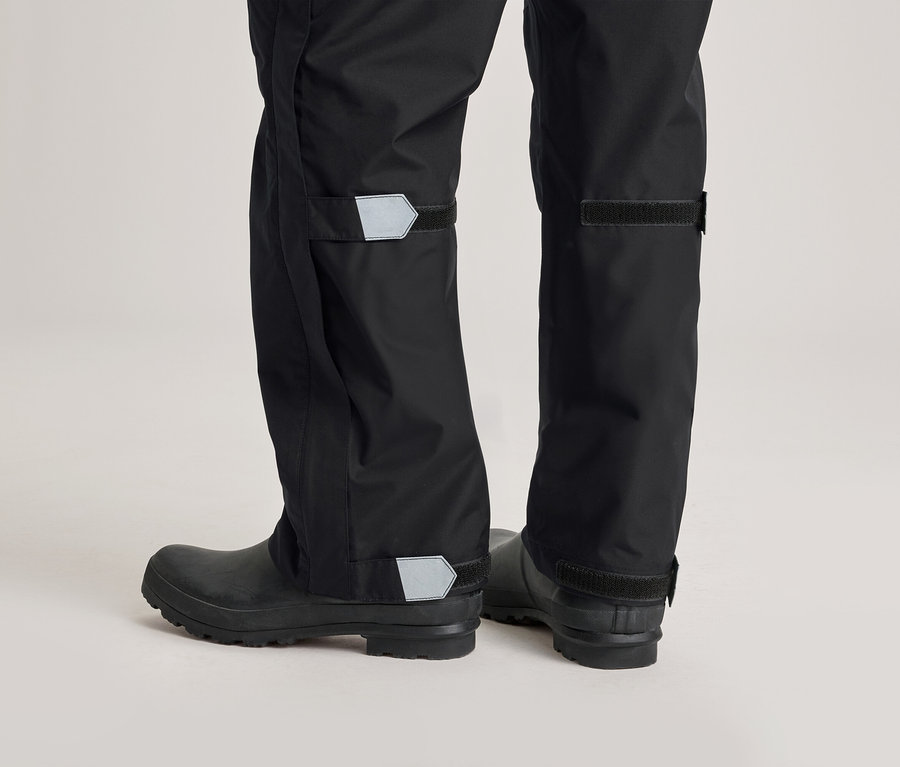 Nahaufnahme der Unterseite einer Person, die schwarze Regenhose und Gummistiefel trägt. Die Hosen haben reflektierende Details.