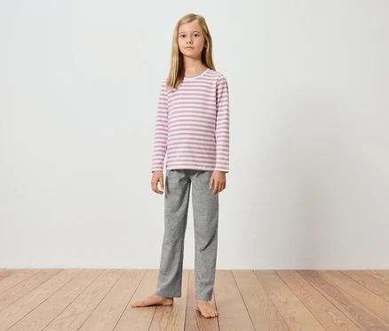 Ein junges Mädchen steht barfuß auf einem Holzboden vor einer weißen Wand. Sie trägt ein langärmeliges, rosa-weiß gestreiftes Oberteil und eine graue Hose.