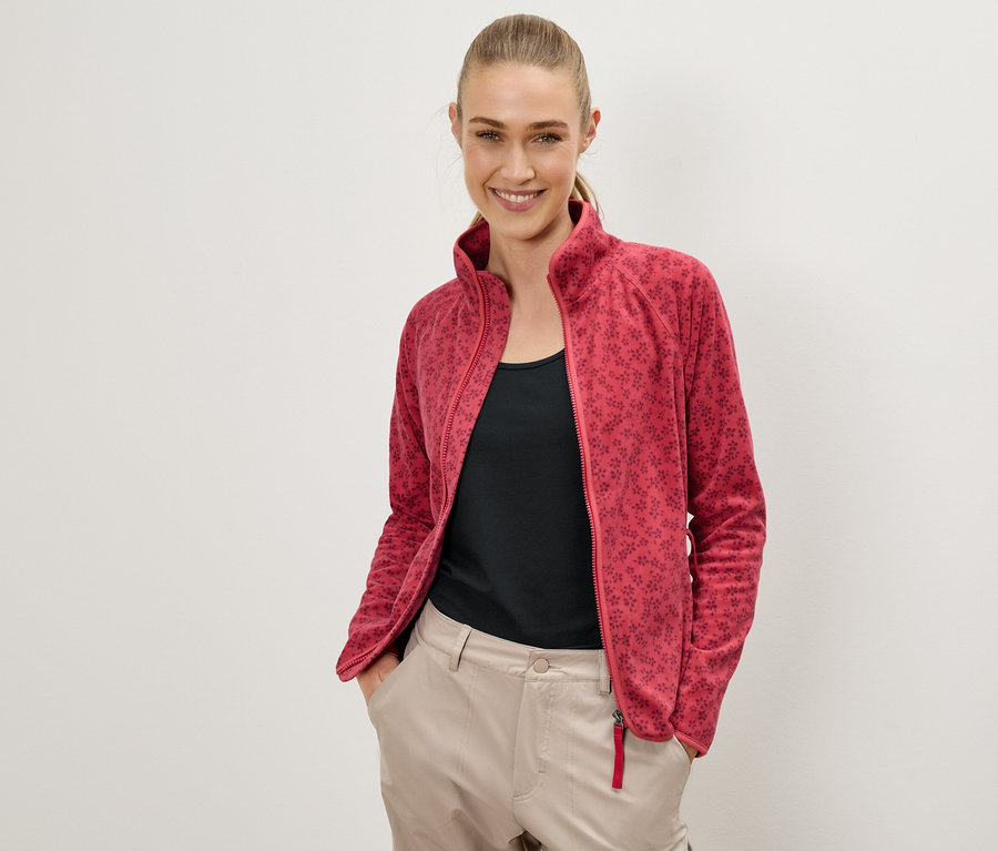 Frau mit einer roten Fleecejacke mit Blumenmuster, einem schwarzen Top und beigen Hosen, die vor einer weißen Wand posiert und lächelt.