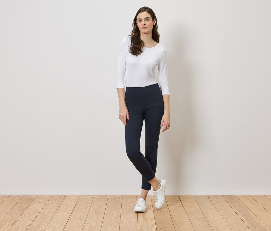 Frau steht vor einer weißen Wand, gekleidet in ein weißes Oberteil, blaue Leggings und weiße Turnschuhe.