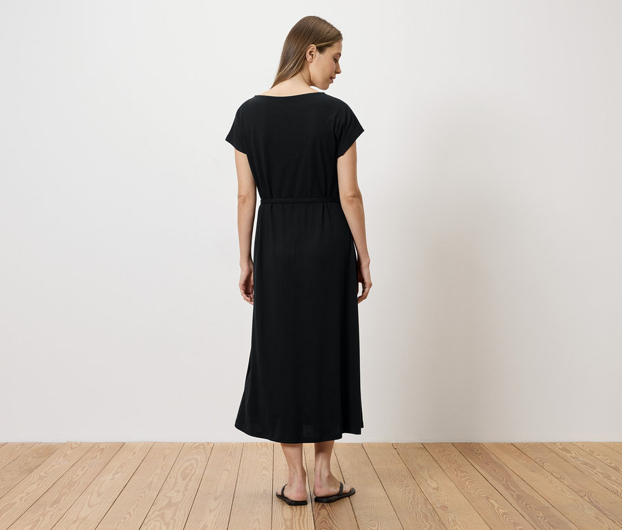 Eine Frau steht mit dem Rücken zur Kamera und trägt ein schwarzes, knöchellanges Kleid mit Gürtel und flache Sandalen.