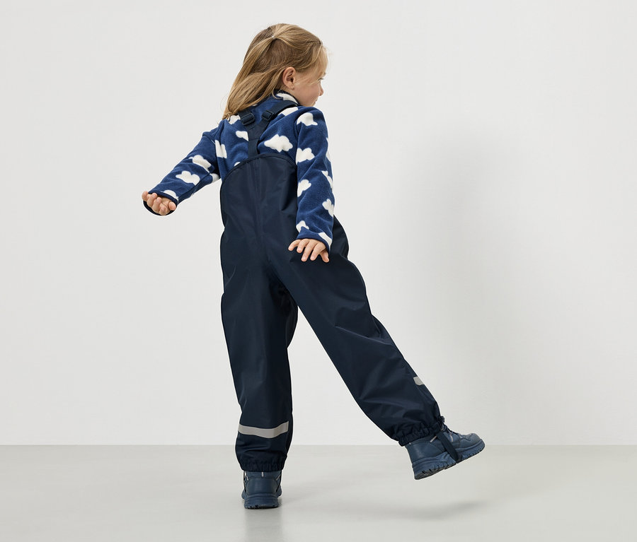 Mädchen in marineblauer Regenhose und blauem Pullover mit Wolkenmuster, von hinten gesehen.