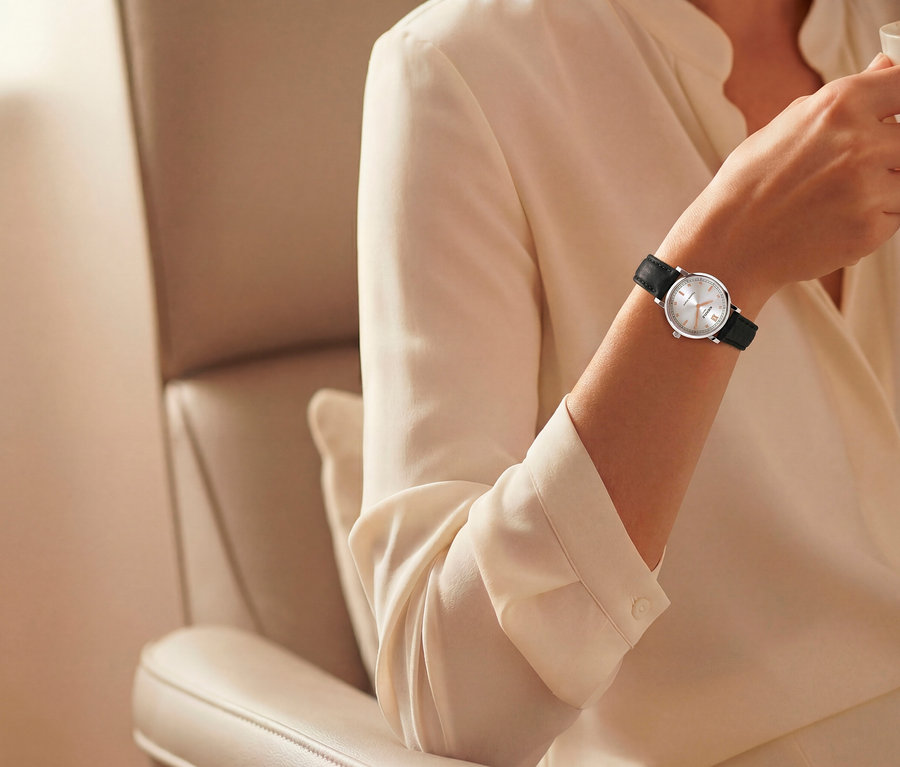 Nahaufnahme eines Arms in einer hellen Bluse, der eine silberne Uhr mit weißem Zifferblatt und schwarzem Armband trägt und ein klares Glas hält, während er in einem beigen Sessel sitzt.