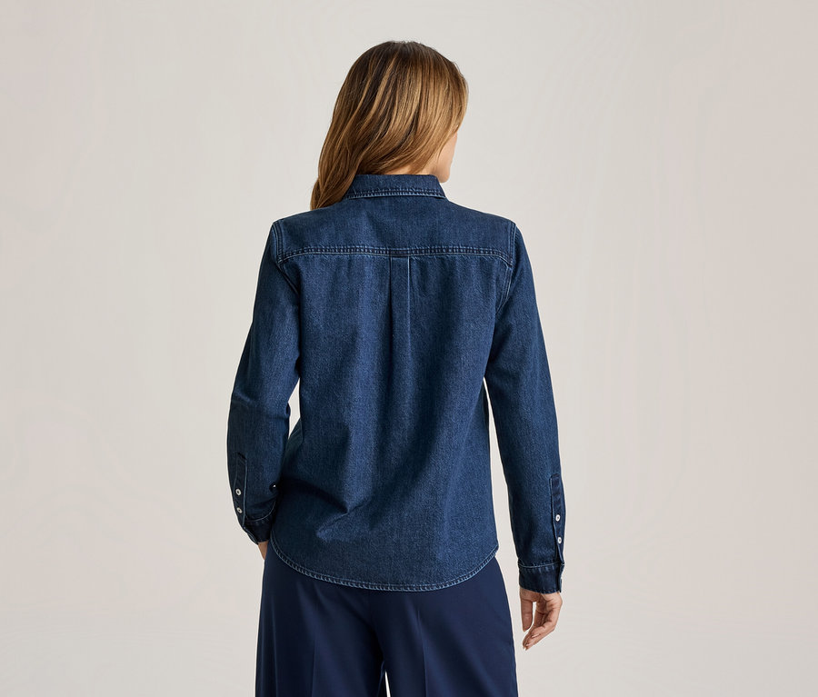Frau von hinten, mit langen, hellbraunen Haaren, trägt ein dunkelblaues Jeanshemd mit langen Ärmeln und eine dunkelblaue weite Hose.