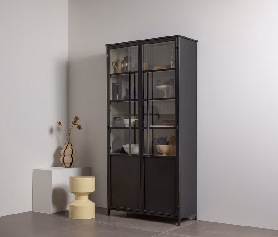 Ein hoher schwarzer Metallvitrinenschrank mit Glastüren oben und massiven Türen unten, gefüllt mit Geschirr und Dekoartikeln, steht in einer weißen Ecke. Links daneben steht ein weißer Sockel mit einer wellenförmigen Vase mit Trockenblumen, daneben ein gelber, sanduhrförmiger Hocker.