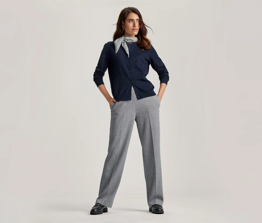 Frau steht mit Händen in den Hosentaschen, trägt einen marineblauen Cardigan, hellgraue weite Hose, einen hellgrauen Schal und schwarze Schuhe.