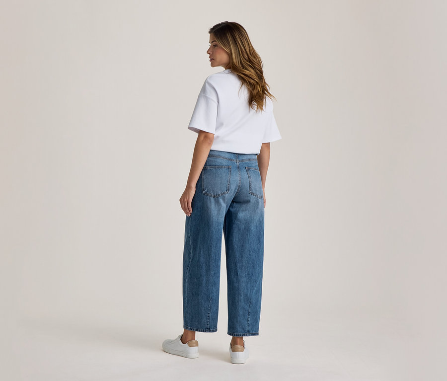 Eine Frau von hinten, leicht gedreht, trägt ein weißes T-Shirt, blaue weite Jeans und weiße Turnschuhe.