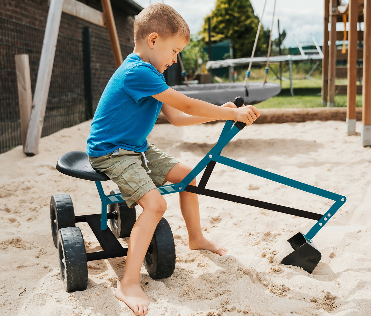Ein kleiner Junge in blauem T-Shirt und Khaki-Shorts sitzt barfuß auf einem small foot Bagger mit Rädern im Sandkasten und bedient dessen Schaufel, um im Sand zu graben.