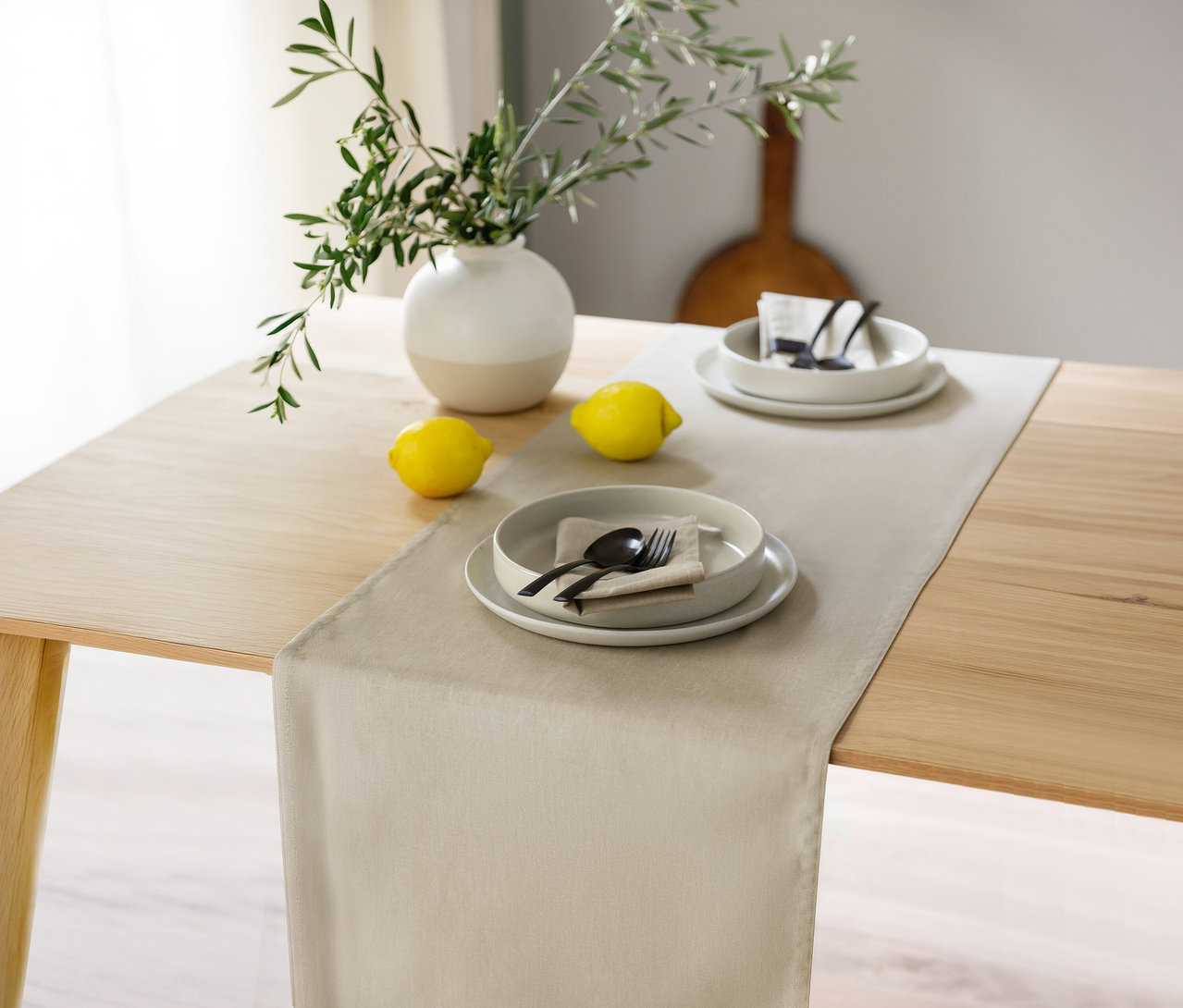 Ein sandfarbener Tischläufer der SCHÖNER WOHNEN-Kollektion liegt auf einem Holztisch. Darauf stehen zwei Gedecke mit Tellern, Schalen, Servietten und schwarzem Besteck, flankiert von zwei Zitronen und einer Vase mit Olivenzweigen.