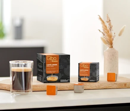 Zwei Qbo Caffè MINAS VERDE Kapselpackungen, ein Glas Kaffee und lose Kapseln auf einem Holzbrett auf der Küchenzeile, daneben Kekse und Trockenblumen.