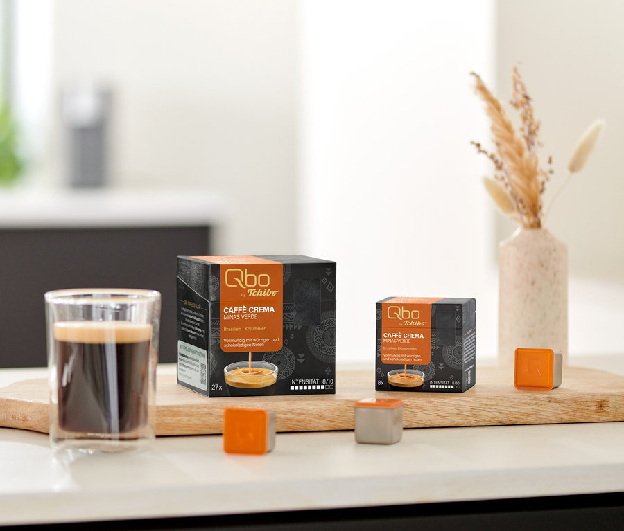 Zwei Qbo Caffè MINAS VERDE Kapselpackungen, ein Glas Kaffee und lose Kapseln auf einem Holzbrett auf der Küchenzeile, daneben Kekse und Trockenblumen.