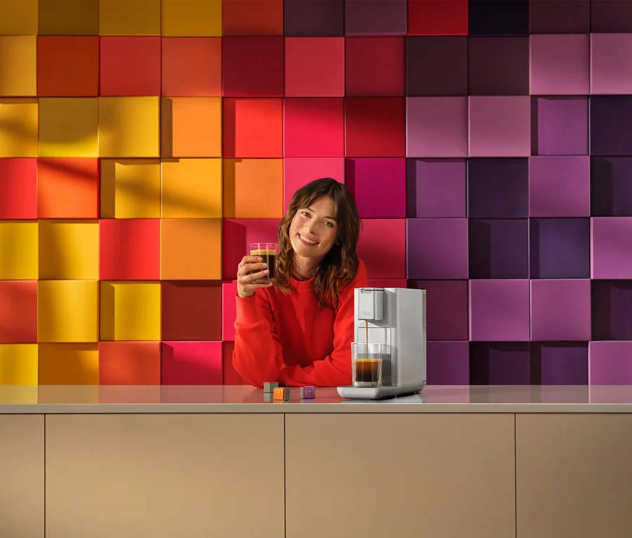 Lächelnde Frau in rotem Oberteil hält ein Glas Kaffee, während eine silberne Kaffeemaschine Kaffee in ein Glas füllt. Bunter Hintergrund aus quadratischen Blöcken.