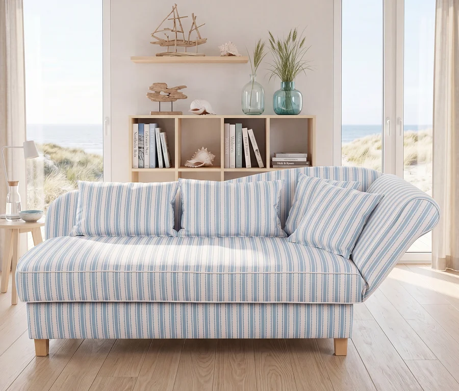 Ein helles, maritim eingerichtetes Zimmer mit einem hellblau-weiß gestreiften Sofa und passenden Kissen. Dahinter ein Holzregal mit Büchern und maritimer Deko. Große Fenster bieten Meerblick auf Sanddünen.