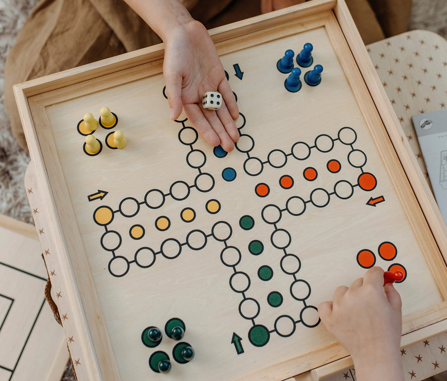 Zwei Personen spielen das Spiel 'Mensch ärgere Dich nicht'. Eine Person hält einen Würfel, die andere bewegt einen roten Spielstein. small foot Spielesammlung, 20 Klassiker.