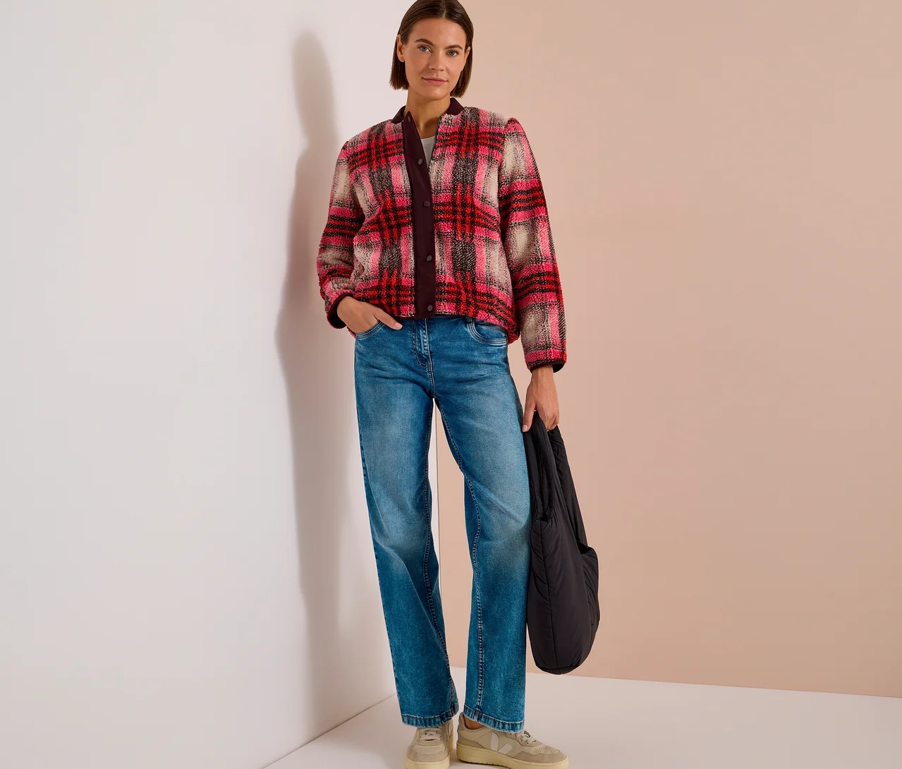 Eine Frau mit kurzen braunen Haaren steht und trägt einen rot-pink-beigen karierten CECIL Blouson, blaue Jeans und helle Sneaker, während sie eine schwarze Tasche hält.