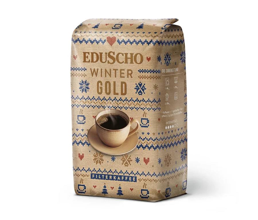 Eine aufrecht stehende Packung EDUSCHO Wiener Gold Ltd. Winter Edition Kaffee mit einem winterlichen Strickmuster in Beige, Blau und Rot, verziert mit Schneeflocken, Herzen und Kaffeetassen. Auf der Vorderseite ist eine dampfende Tasse Kaffee abgebildet.