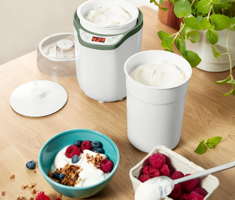 Ein weißer Joghurt-und-Frischkäsebereiter mit grünen Akzenten steht auf einem Holztisch. Er enthält Joghurt, ebenso wie ein separater Becher und eine türkise Schale mit Joghurt, Beeren und Müsli. Daneben ist eine Schale mit Himbeeren und einem Löffel.