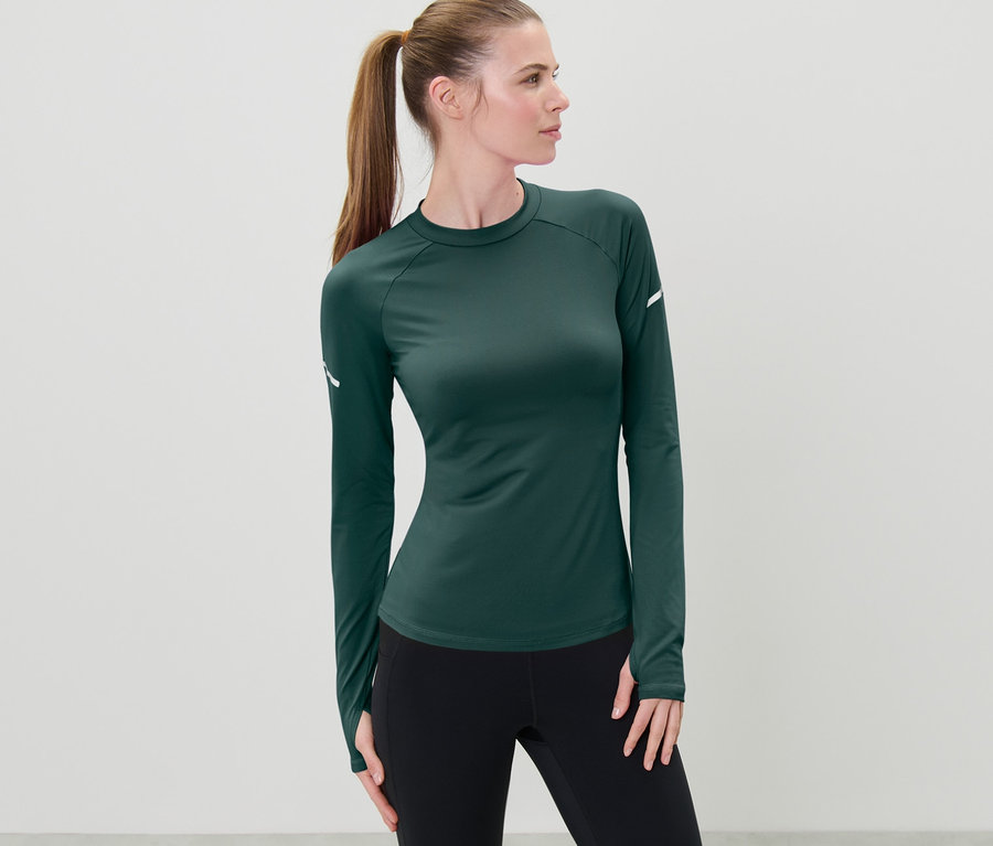 Frau mit braunen Haaren, die zu einem Pferdeschwanz zusammengebunden sind, posiert in einem dunkelgrünen Langarm-Funktionsshirt.