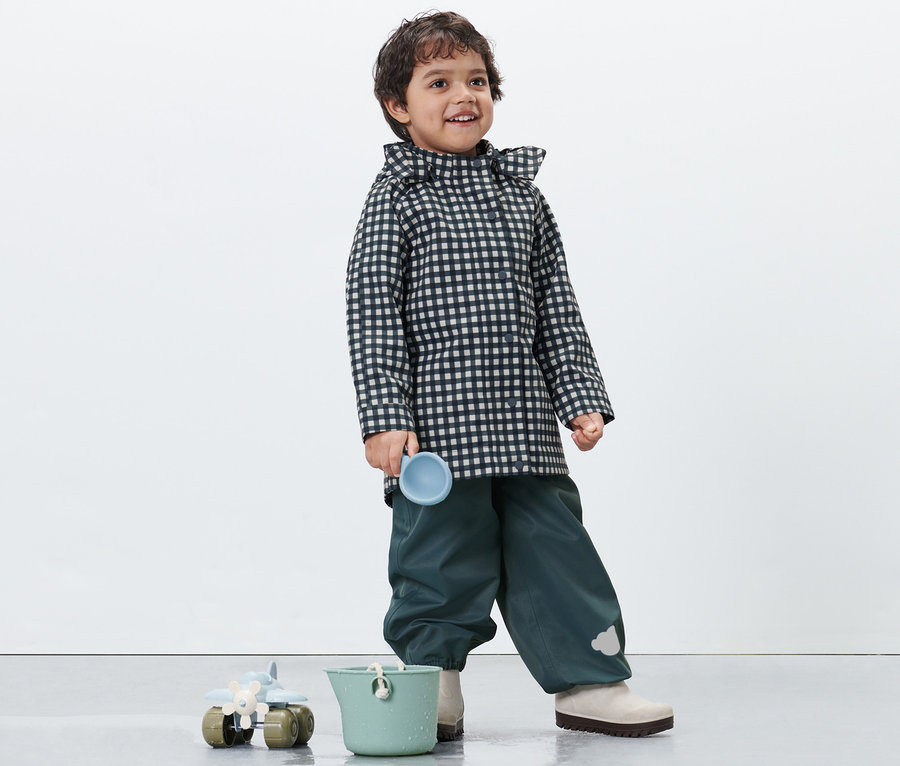 Ein Junge in einer karierten Kleinkind-Regenjacke hält eine blaue Tasse. Unten stehen ein grüner Eimer und ein Spielzeug.