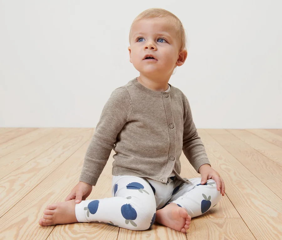 Baby sitzt auf einem Holzboden und trägt eine Baby-Strickjacke.