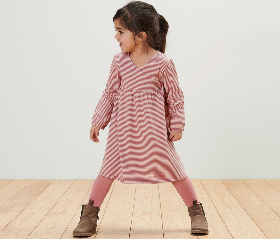 Mädchen steht auf einem Holzboden und trägt ein Kinder-Jerseykleid mit rosa Leggings und braunen Stiefeln.