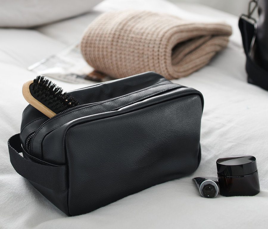 Eine schwarze Kulturtasche mit Haarbürste, Creme und Flasche auf einem weißen Bettlaken.
