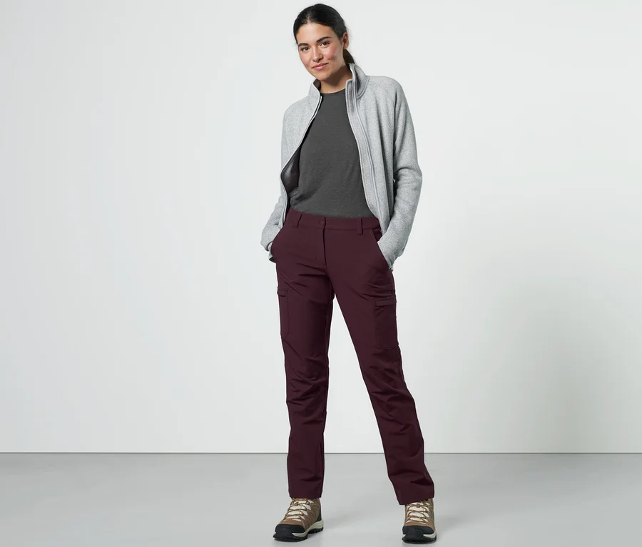 Eine Frau posiert in einer bordeauxroten Cargo-Softshellhose, einem grauen T-Shirt und einer Sweatshirtjacke.