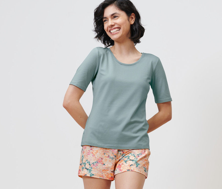 Frau steht und lächelt, sie trägt einen Shorty-Pyjama, Shorts mit Blumen-Alloverprint und ein T-Shirt.