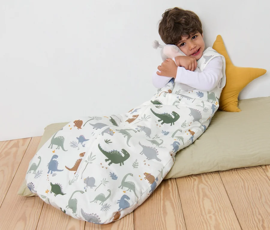Junge liegt in einem Schlafsack 3 TOG, 110 cm mit Dinosaurier-Muster und umarmt ein Stofftier.