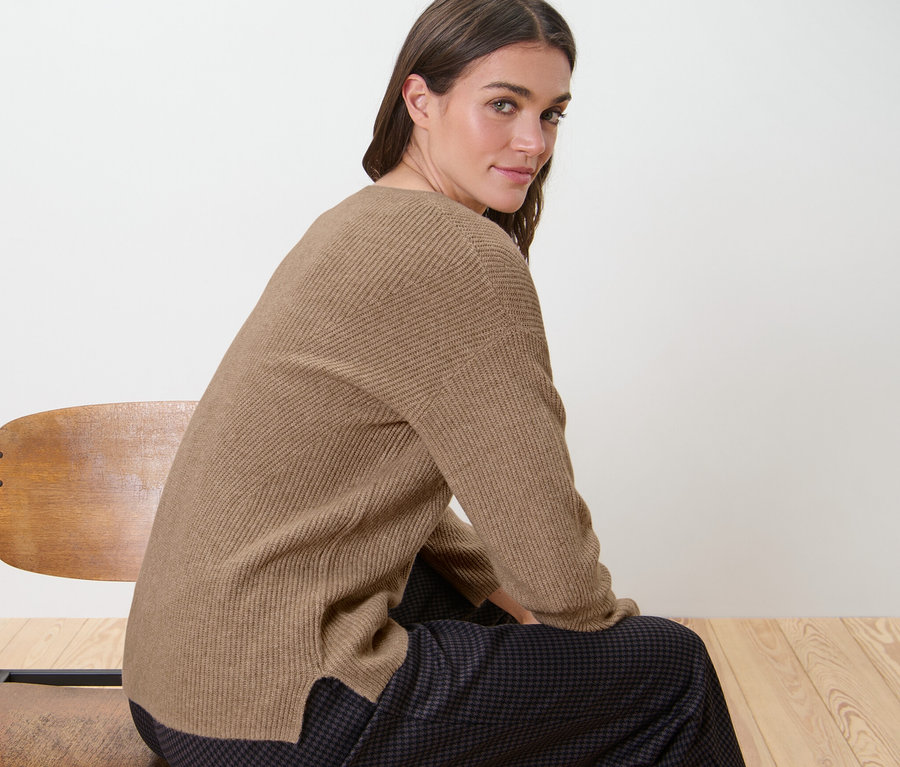 Frau sitzt auf einem Holzstuhl und trägt einen kamelfarbenen Grobstrickpullover.