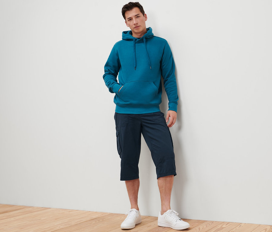 Mann lehnt an Wand, trägt blaugrünen Hoodie und navy Cargo-Bermuda in 3/4-Länge.