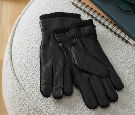 Schwarze Lederhandschuhe liegen auf einem beige Hocker neben einem Buch und einer Brille.