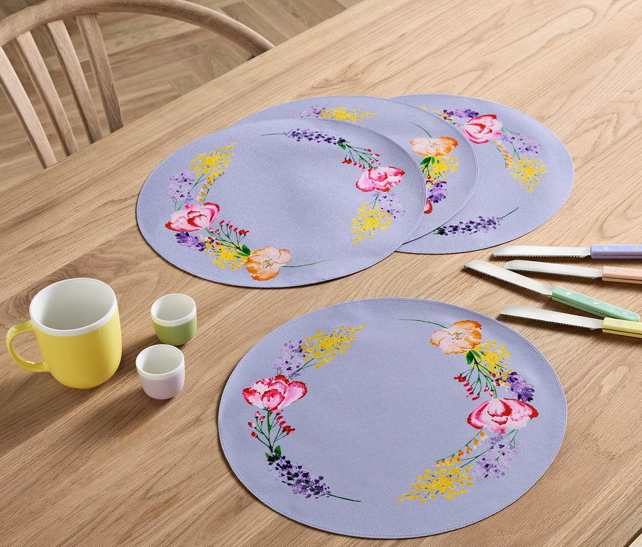 Auf einem Holztisch liegen vier Platzsets mit Blumenmuster, eine gelbe Tasse, zwei kleine Tassen und vier Messer mit bunten Griffen.