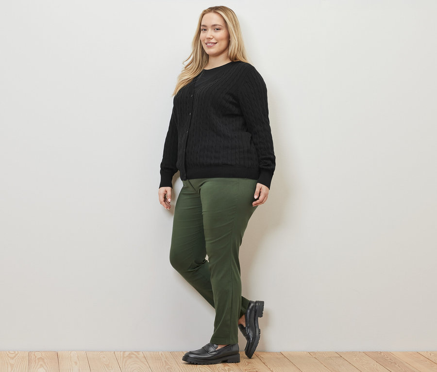 Frau posiert in Feinstrickcardigan mit Zopfmuster in Schwarz und einer Bengalin-Hose in Grün.