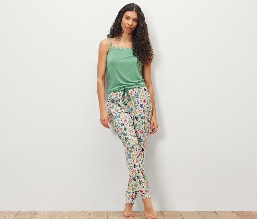 Eine Frau steht vor einer weißen Wand und trägt ein grünes Top und eine Pyjamahose.