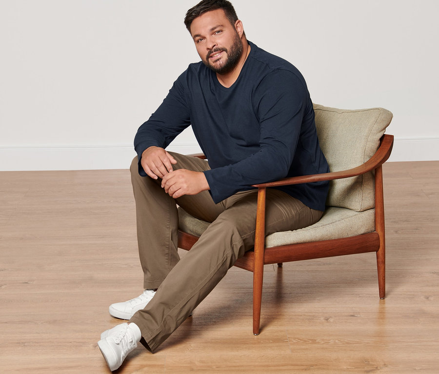 Ein Mann sitzt in einem Sessel und trägt 2 Langarmshirts, eine sandfarbene Chino und Sneaker.