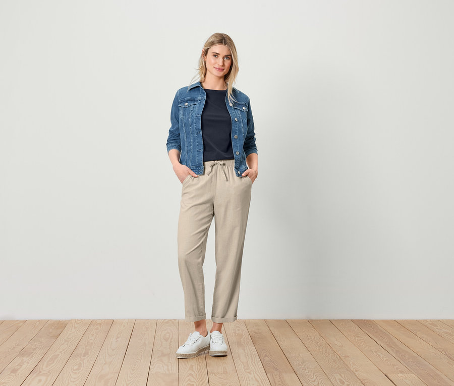 Eine Frau posiert in Jeansjacke, sandfarbener Leinenhose und einem dunkelblauen Basic T-Shirt.