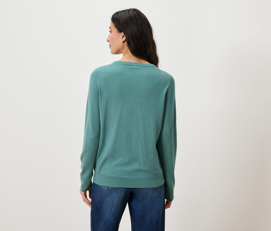 Rückansicht einer Frau mit langen, dunklen Haaren, die blaue Jeans und einen grünen Pullover trägt.