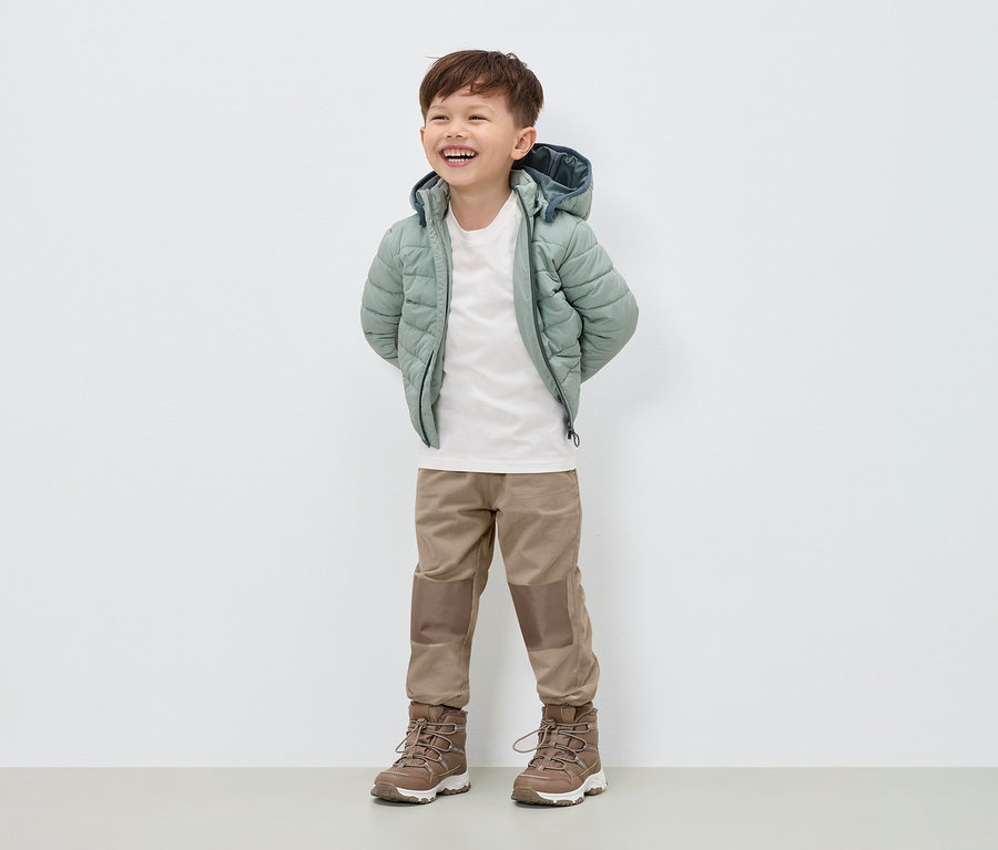 Ein lächelnder Junge steht in einer hellgrünen Daunenjacke, beigen Hosen und braunen Stiefeln.