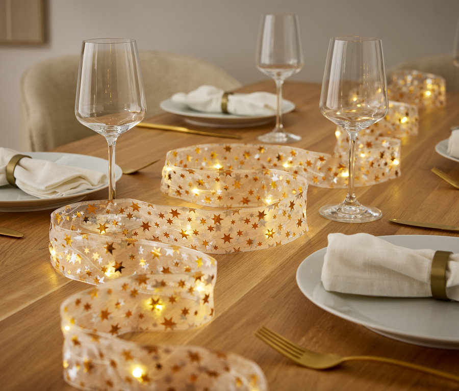 Ein gedeckter Tisch mit Tellern, Weingläsern, goldenem Besteck und einer Lichterkette mit goldenen Sternen.