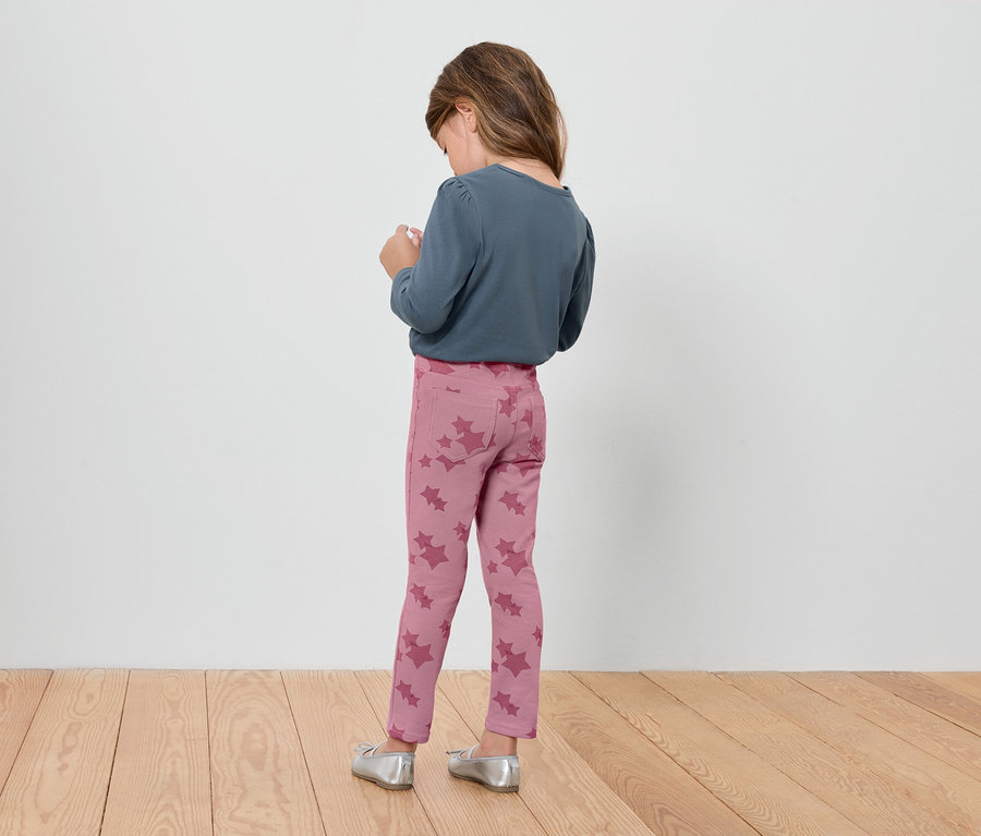 Mädchen in rosa Leggings mit Blumenmuster und blauem Langarmshirt steht mit dem Rücken zum Betrachter.