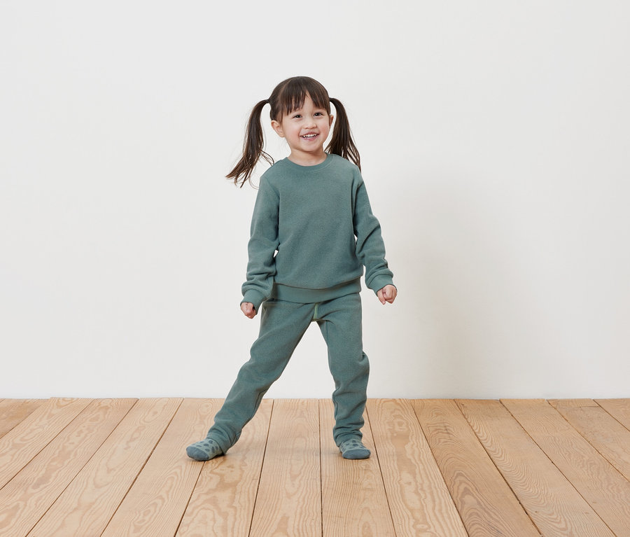 Ein lächelndes kleines Mädchen mit Zöpfen posiert in einem hellgrünen Sweatshirt- und Jogginghosen-Set auf einem Holzboden vor einer weißen Wand.