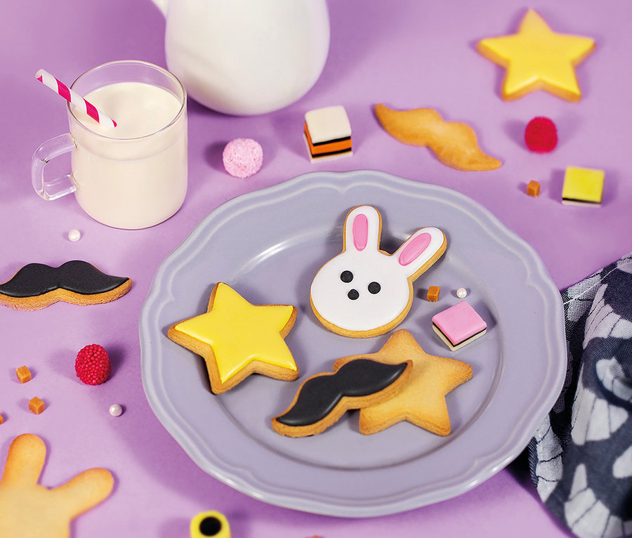 Teller mit stern-, hasen- und schnurrbartförmigen Keksen, neben einem Glas Milch, einer Kanne und Süßigkeiten auf violettem Hintergrund. Chefclub Dessert und Keks-Back-Set.