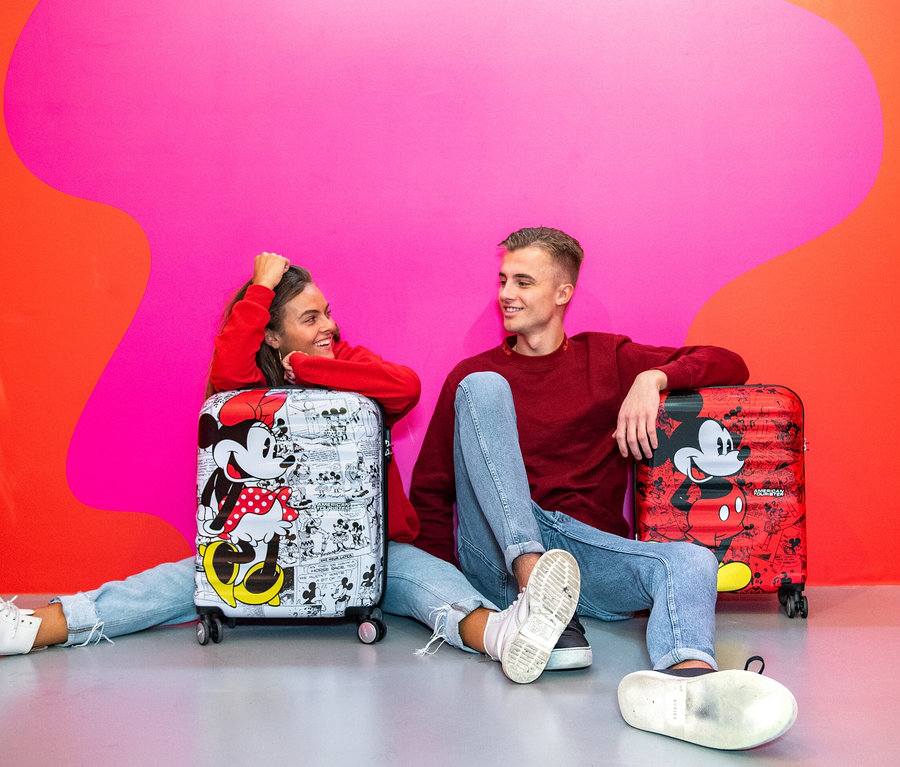 Zwei junge Leute sitzen vor American Tourister Wavebreaker Koffern mit Mickey und Minnie Mouse Motiv vor einer farbigen Wand.