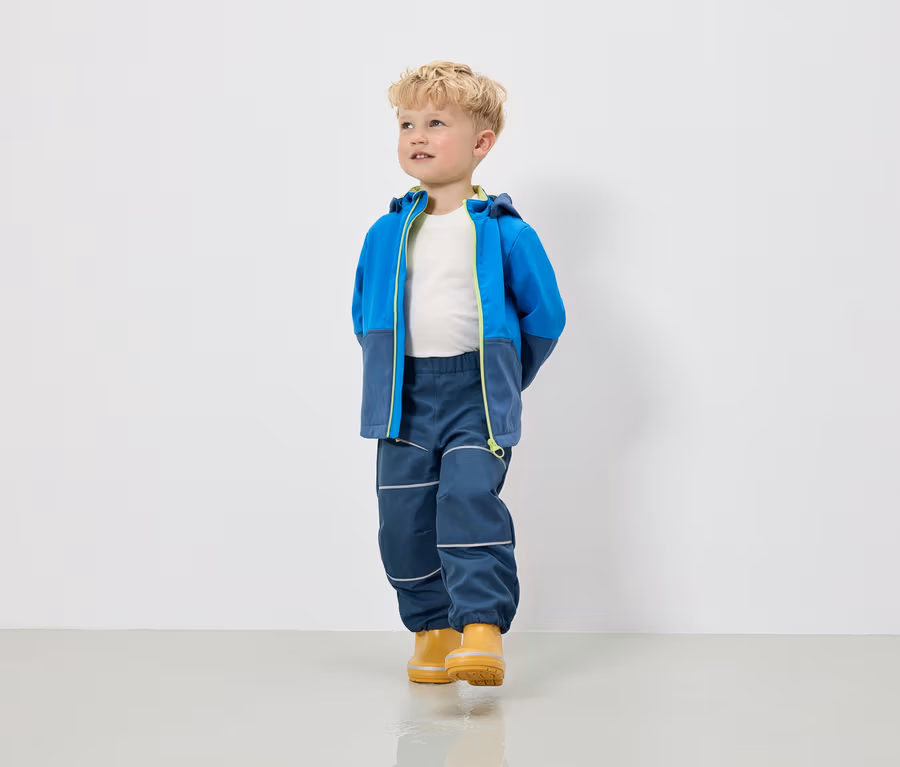 Ein Junge in einer blau-grauen Jacke mit Kapuze, Hose und gelben Gummistiefeln steht vor einer weißen Wand.