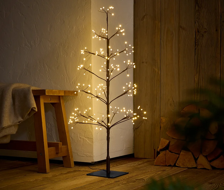 Ein dekorativer Baum mit warmweißen Lichtern steht auf einem Holzboden neben einer Bank.