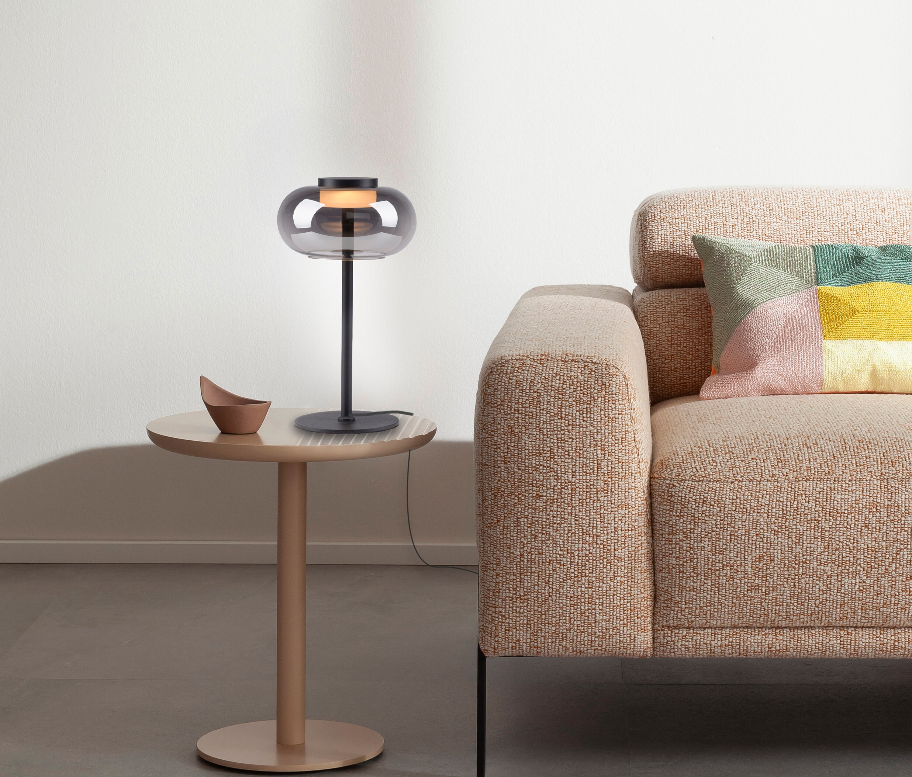 Graue Lampe steht auf rundem Tisch neben beigem Sofa mit buntem Kissen.